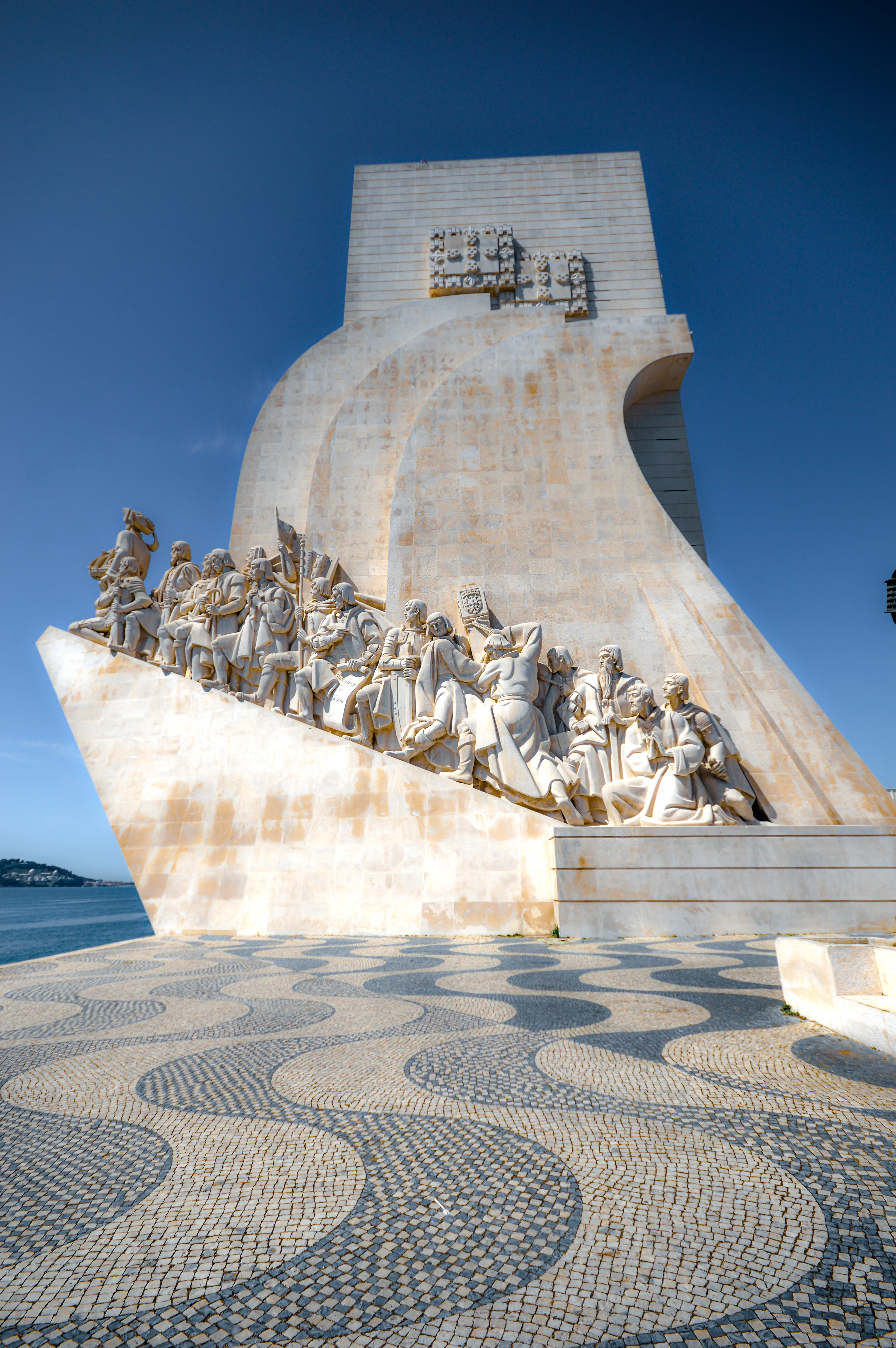 The Monument to the Discoveries in Lisbon, Portugal, featuring carved figures on a ship's prow-shaped structure, under a bright blue sky. The foreground shows a patterned cobblestone walkway.